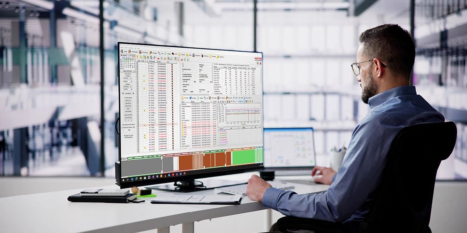 A man in an office setting works on a large computer monitor displaying complex data and graphs, with a tablet beside him.