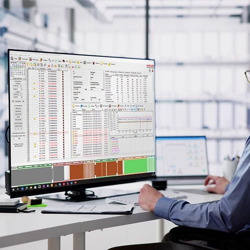 A man in an office setting works on a large computer monitor displaying complex data and graphs, with a tablet beside him.