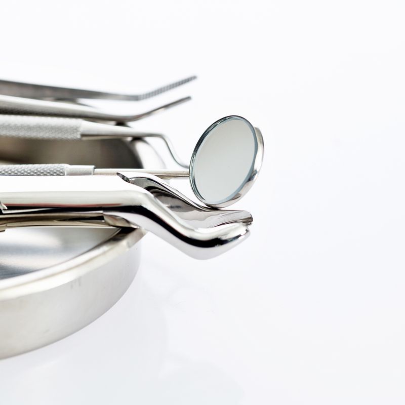 A set of shiny dental tools, including a mirror and tweezers, resting on a metal tray against a white background.