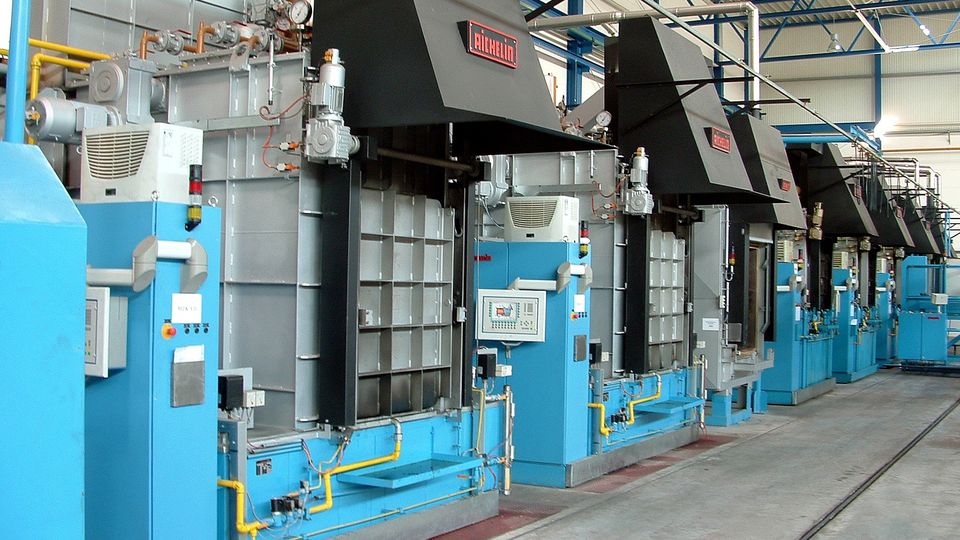 A row of industrial chamber furnaces with blue bases and black hoods lined up in a spacious factory setting with a high, metal ceiling.
