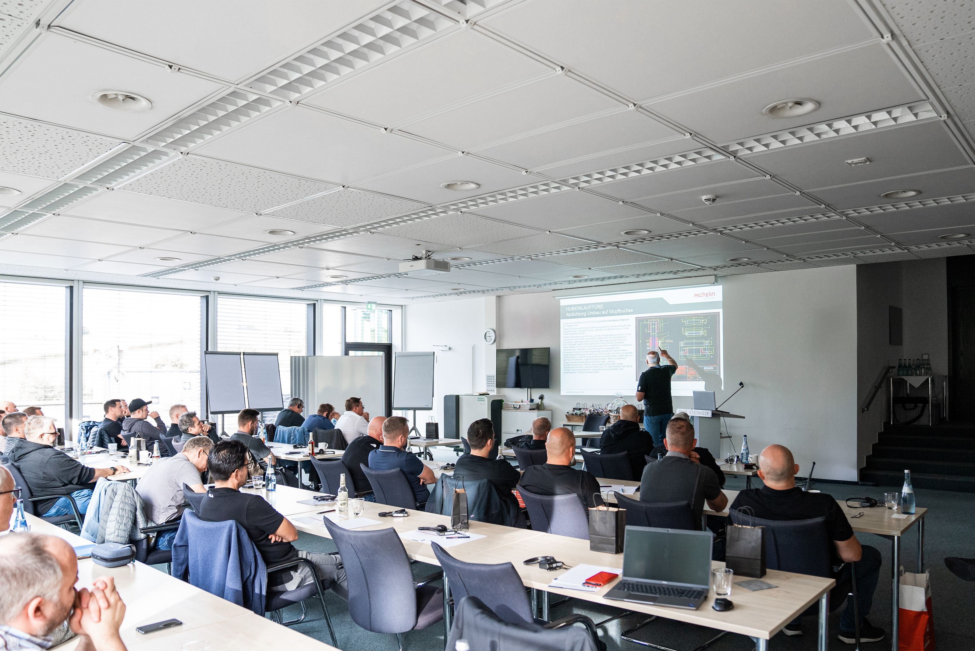 A group of people in a classroom setting listening to a presentation with slides projected on a screen. Laptops and notepads are on tables.