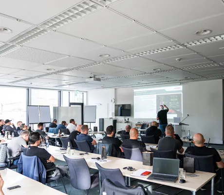A group of people in a classroom setting listening to a presentation with slides projected on a screen. Laptops and notepads are on tables.
