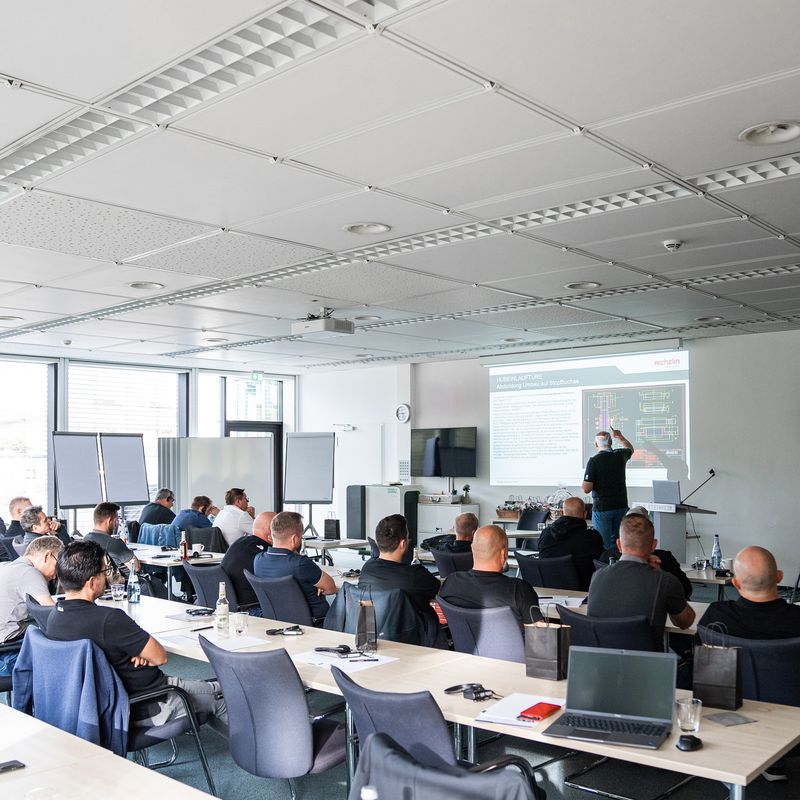 A group of people in a classroom setting listening to a presentation with slides projected on a screen. Laptops and notepads are on tables.
